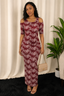 A woman modeling a burgundy and white floral print cold-shoulder maxi dress, holding a beige quilted handbag, set against a cream curtain backdrop with a potted plant.