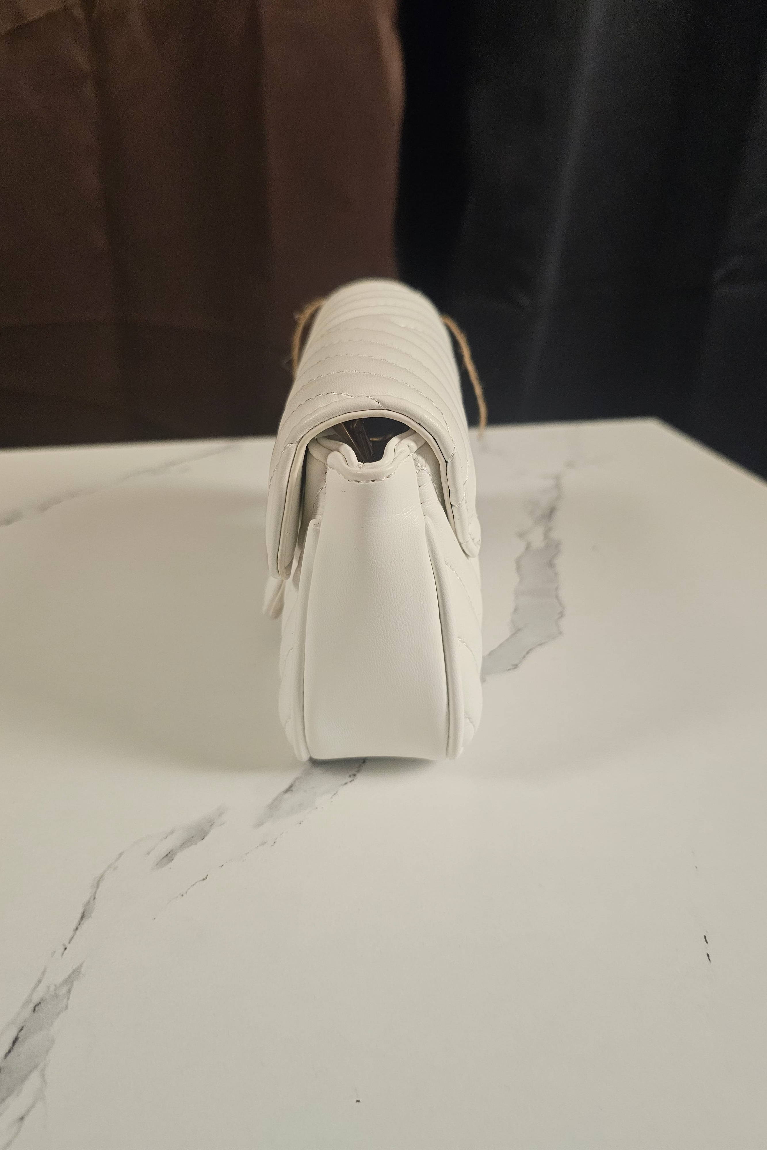 A white faux-leather shoulder bag with a quilted texture and a light brown leather strap, shown from a rear three-quarter angle on a white marble surface.