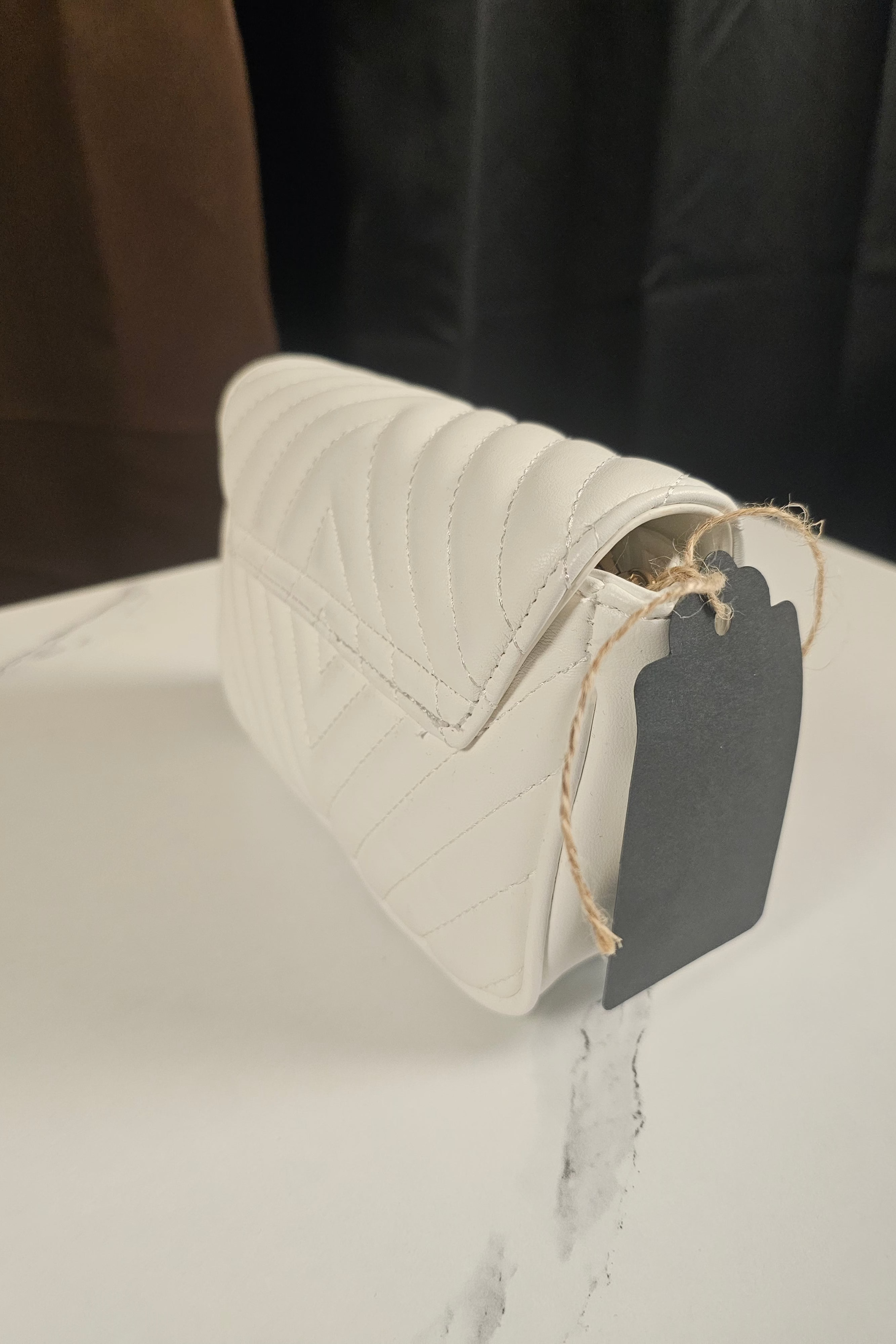 A white quilted leather shoulder bag with a flap closure, resting on a white marble surface with a black fabric backdrop. A black paper tag is attached to the bag with a piece of twine.
