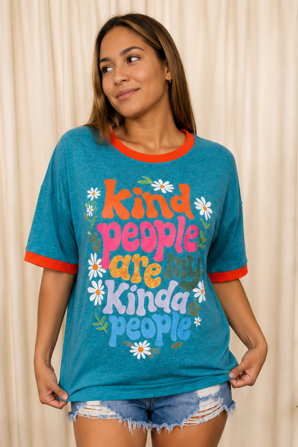A woman wearing a heather teal graphic t-shirt with orange trim, featuring the retro-style text 'kind people are kinda people' surrounded by white daisies, paired with distressed denim shorts against a beige curtain backdrop.