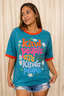 A woman wearing a heather teal graphic t-shirt with orange trim, featuring the retro-style text 'kind people are kinda people' surrounded by white daisies, paired with distressed denim shorts against a beige curtain backdrop.