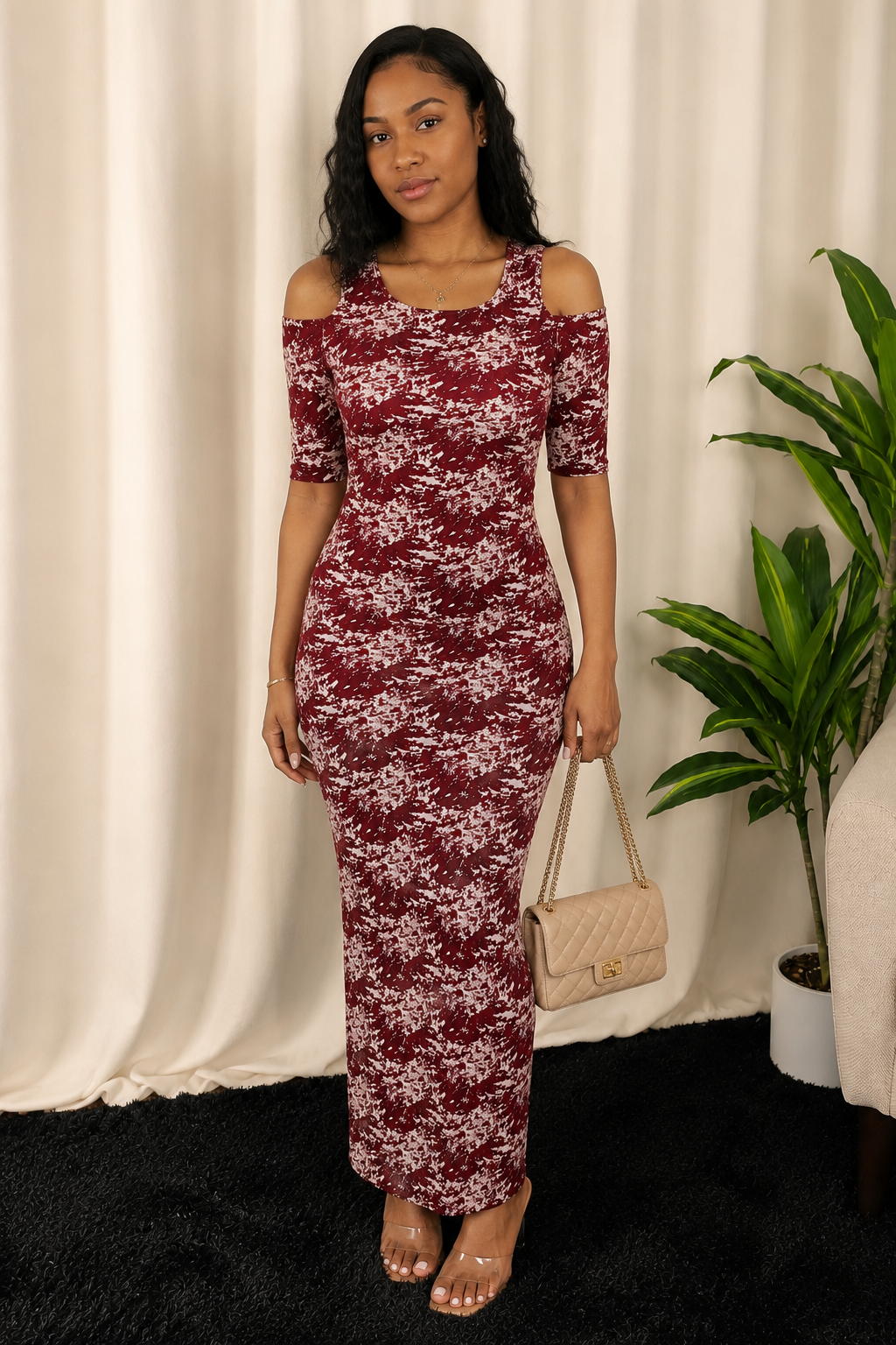 A woman modeling a burgundy and white floral print cold-shoulder maxi dress, holding a beige quilted handbag, set against a cream curtain backdrop with a potted plant.