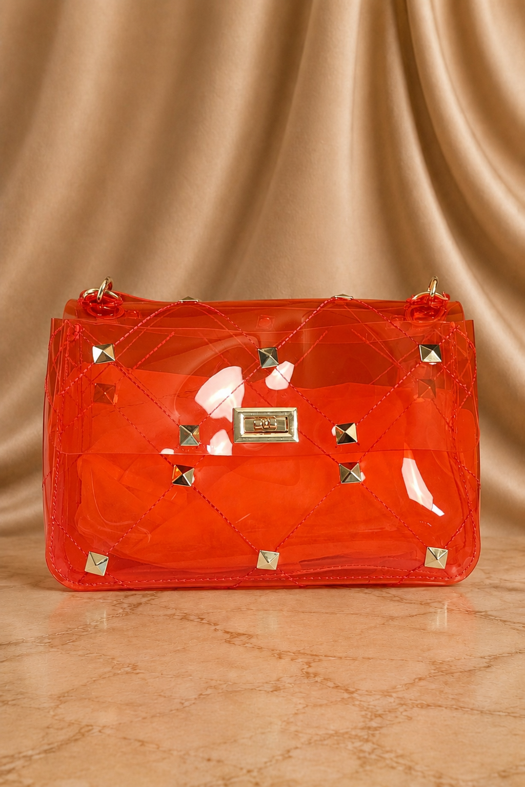A translucent orange-red handbag with a quilted pattern, gold-tone studs, and a central metal plaque, set against a draped beige satin background.