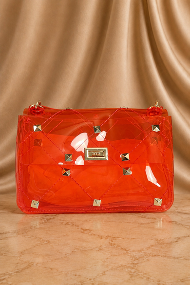A translucent orange-red handbag with a quilted pattern, gold-tone studs, and a central metal plaque, set against a draped beige satin background.