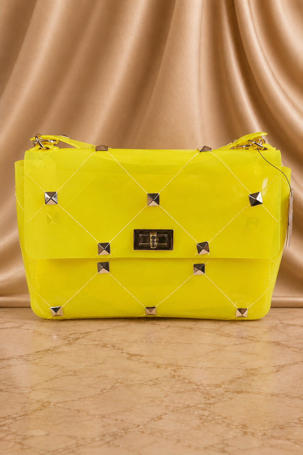 A bright yellow quilted shoulder bag with silver pyramid studs and a black metal clasp, set against a draped beige satin background.