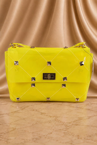 A bright yellow quilted shoulder bag with silver pyramid studs and a black metal clasp, set against a draped beige satin background.