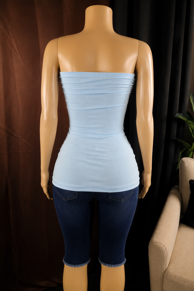 A light blue strapless ruched tube top displayed on a tan mannequin, paired with dark blue capri jeans, set against a dark curtain background.