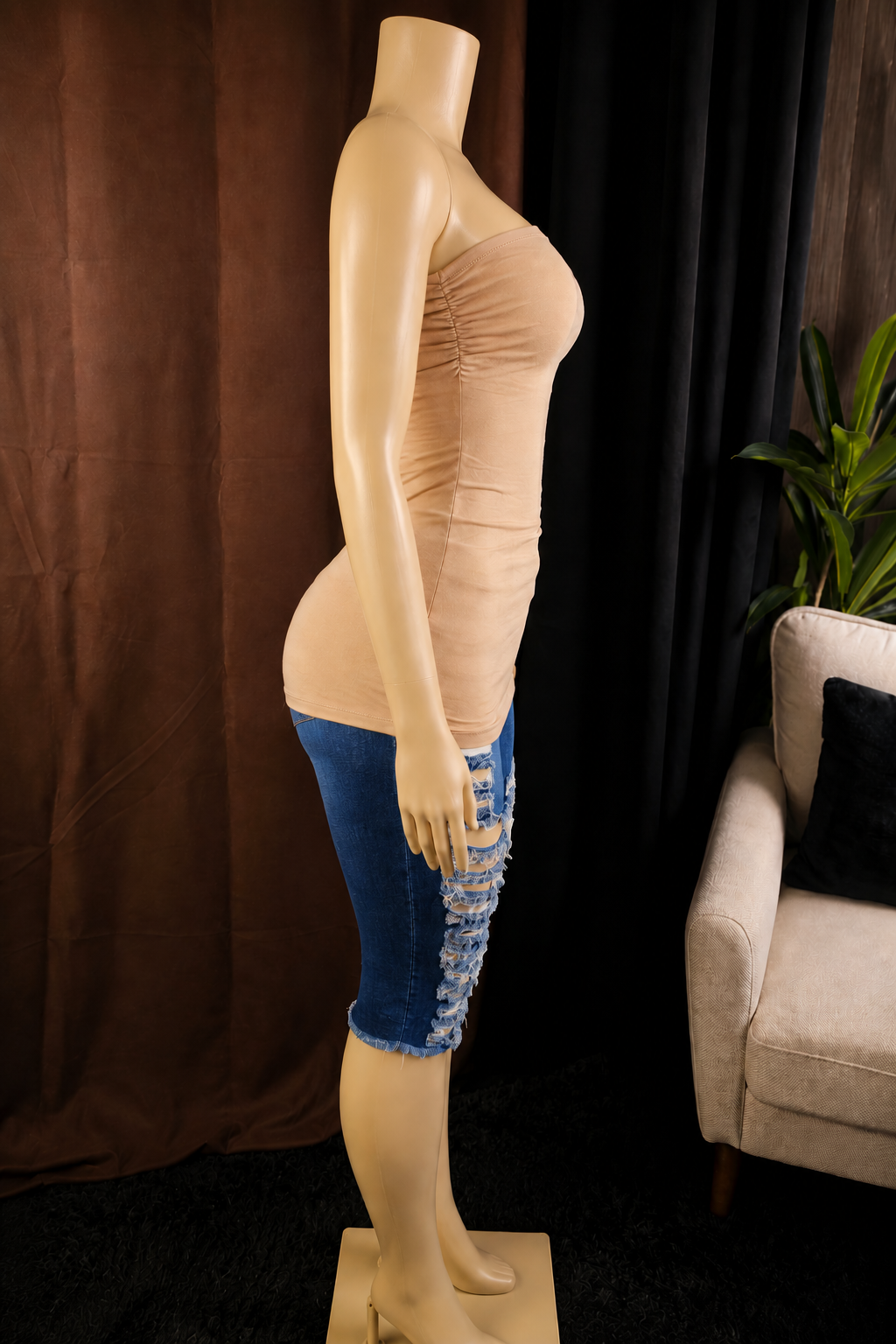 A beige mannequin torso wearing a strapless ruched beige top and blue distressed denim capri pants, shown in profile against a dark curtain backdrop with a chair and plant partially visible.