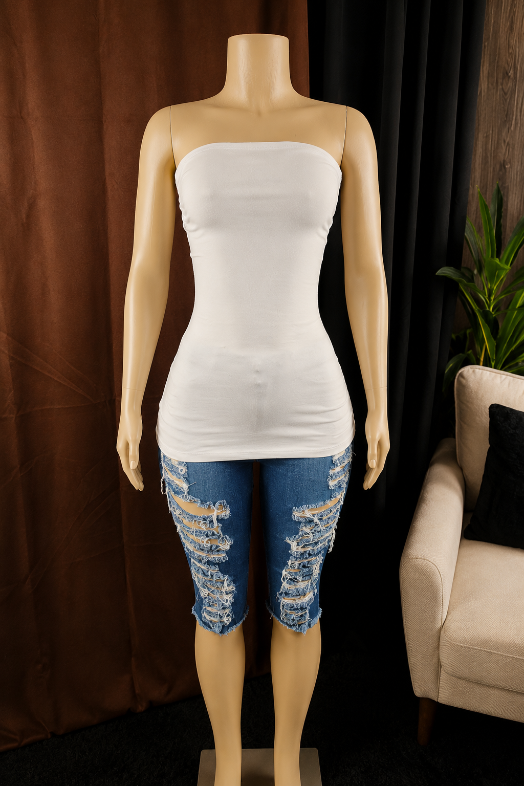 A mannequin wearing a white strapless tube top and distressed blue denim capris, set against a dark brown curtain backdrop with a beige armchair and green plant visible on the right.