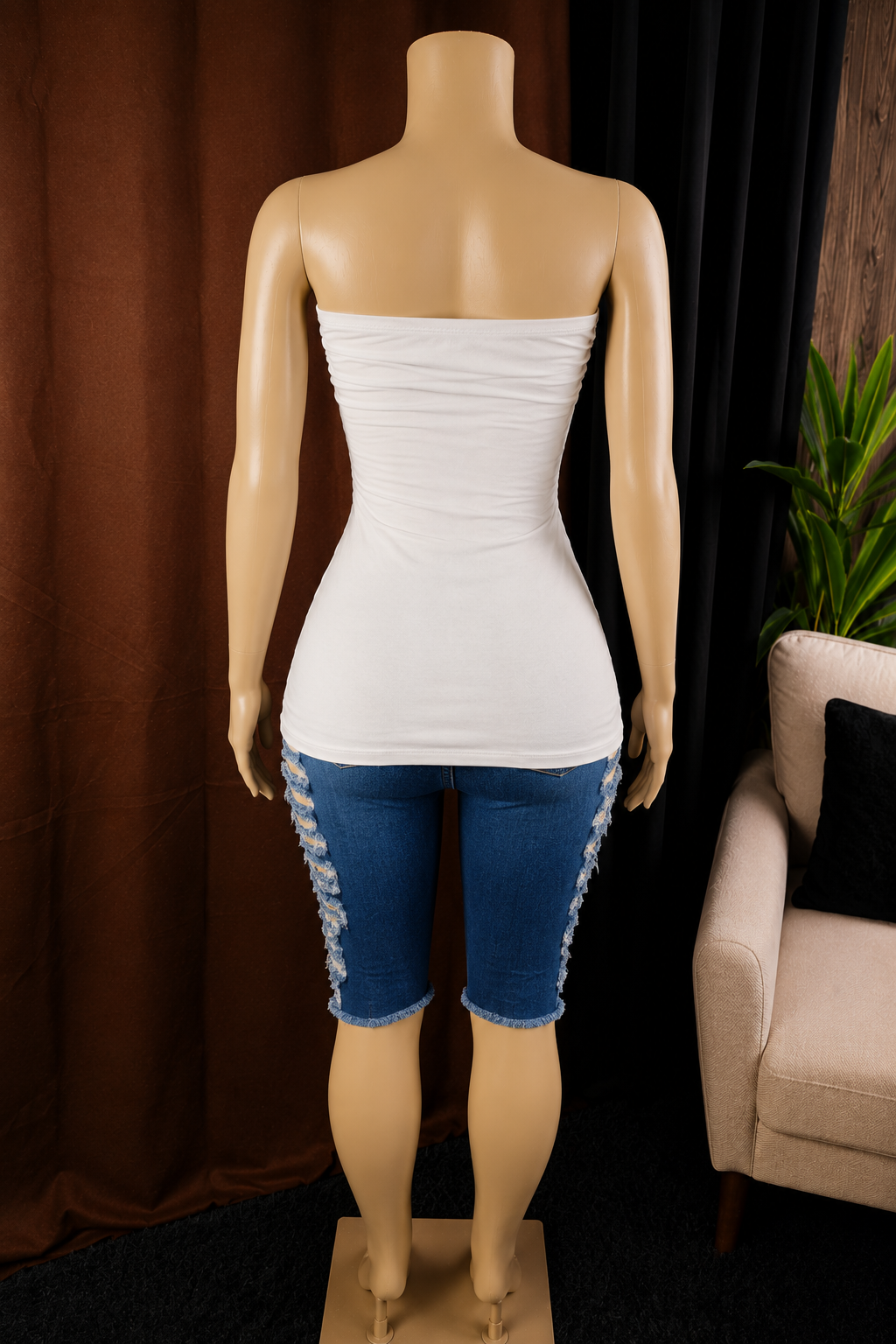 A mannequin wearing a white strapless ruched tube top and blue denim capris with lace-up side details, photographed indoors against dark curtains with a chair and plant partially visible.