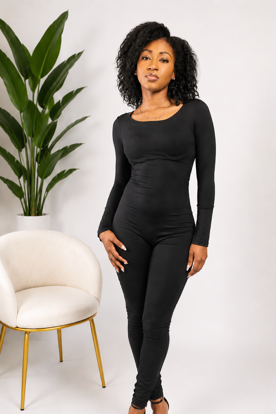 Woman wearing a black jumpsuit standing in a minimalistic room with a plant and chair.