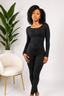 Woman wearing a black jumpsuit standing in a minimalistic room with a plant and chair.