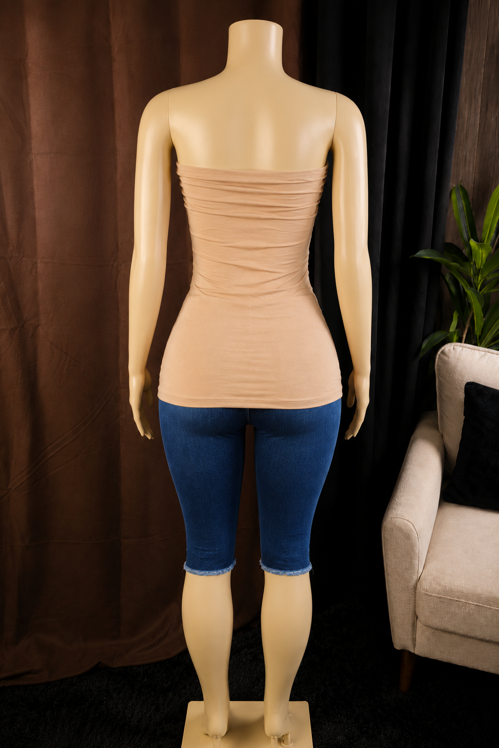 A nude beige strapless ruched tube top displayed on a female mannequin from the back, paired with blue capri jeans, set against a dark curtain backdrop with a chair and plant visible on the right.
