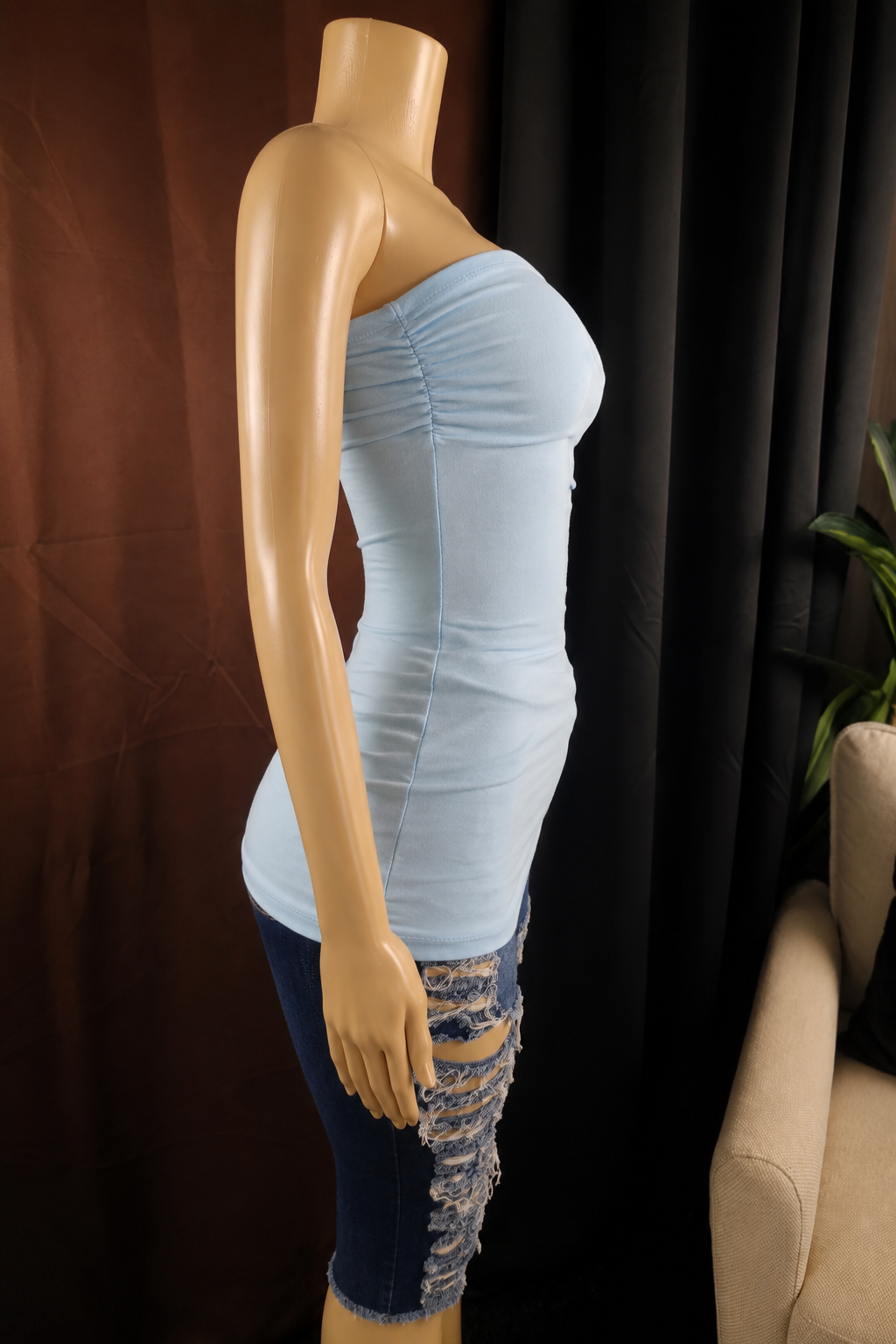 A light blue strapless ruched tube top displayed on a beige mannequin torso, paired with distressed blue jeans, set against a dark brown curtain backdrop.