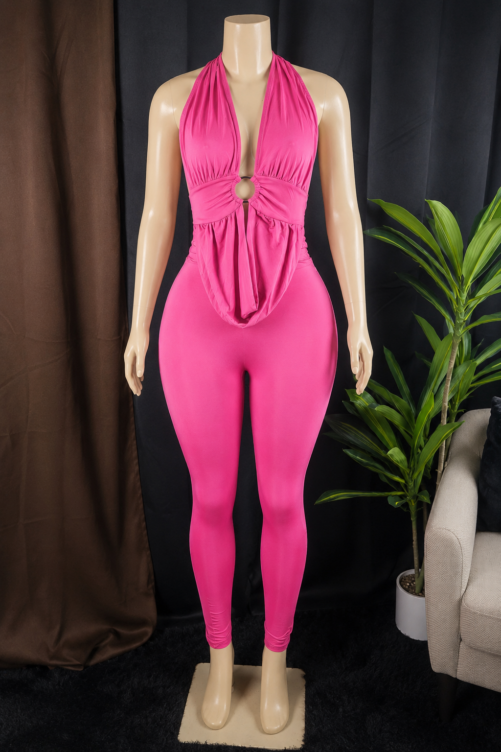 A bright pink one-piece jumpsuit displayed on a beige mannequin, set against a dark backdrop with a brown curtain on the left and a green potted plant on the right.
