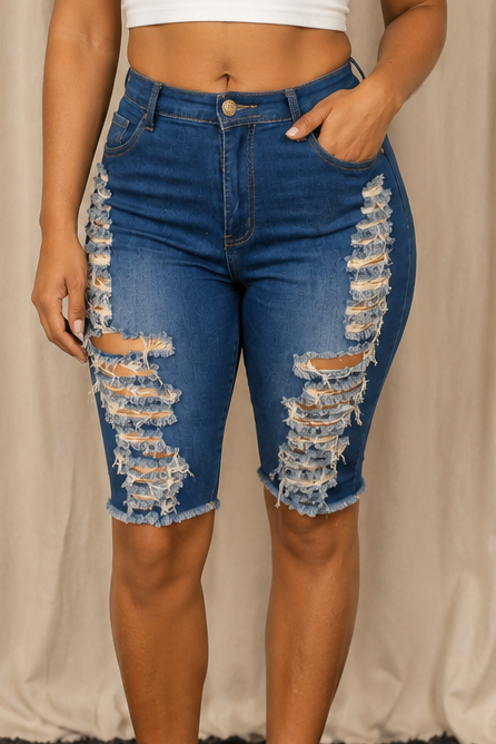 A person wearing medium-wash blue denim cutoff shorts with heavy distressed rips on both legs, paired with a white cropped top, standing against a draped beige fabric backdrop.