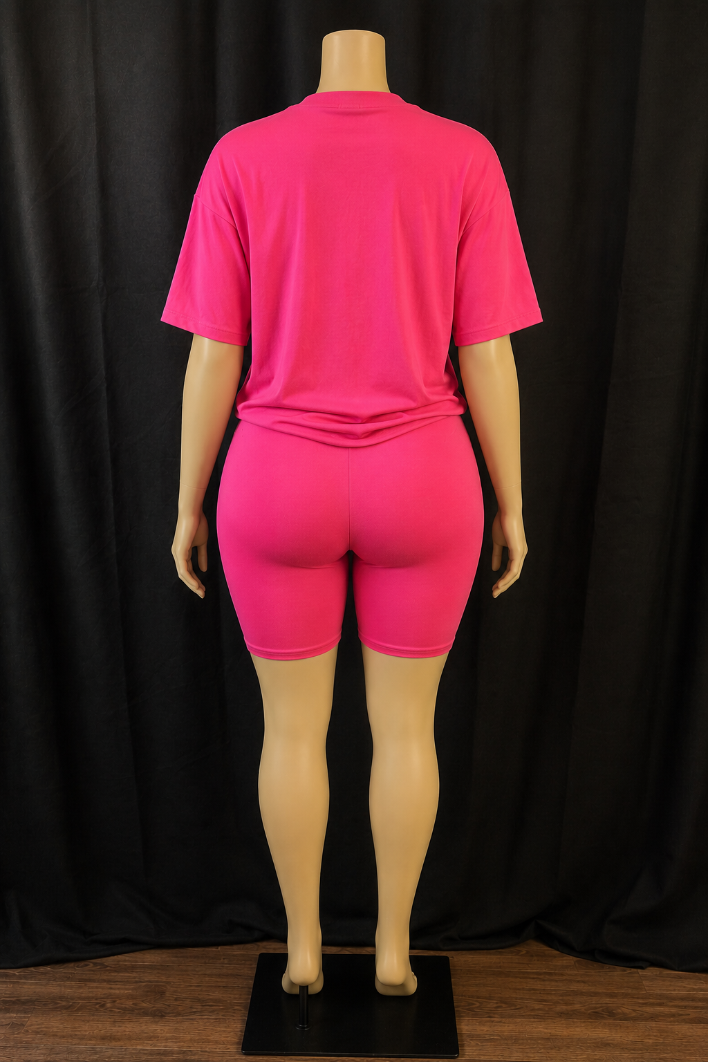 A mannequin viewed from the back wearing a bright pink short-sleeve t-shirt and matching fitted biker shorts against a black curtain backdrop.