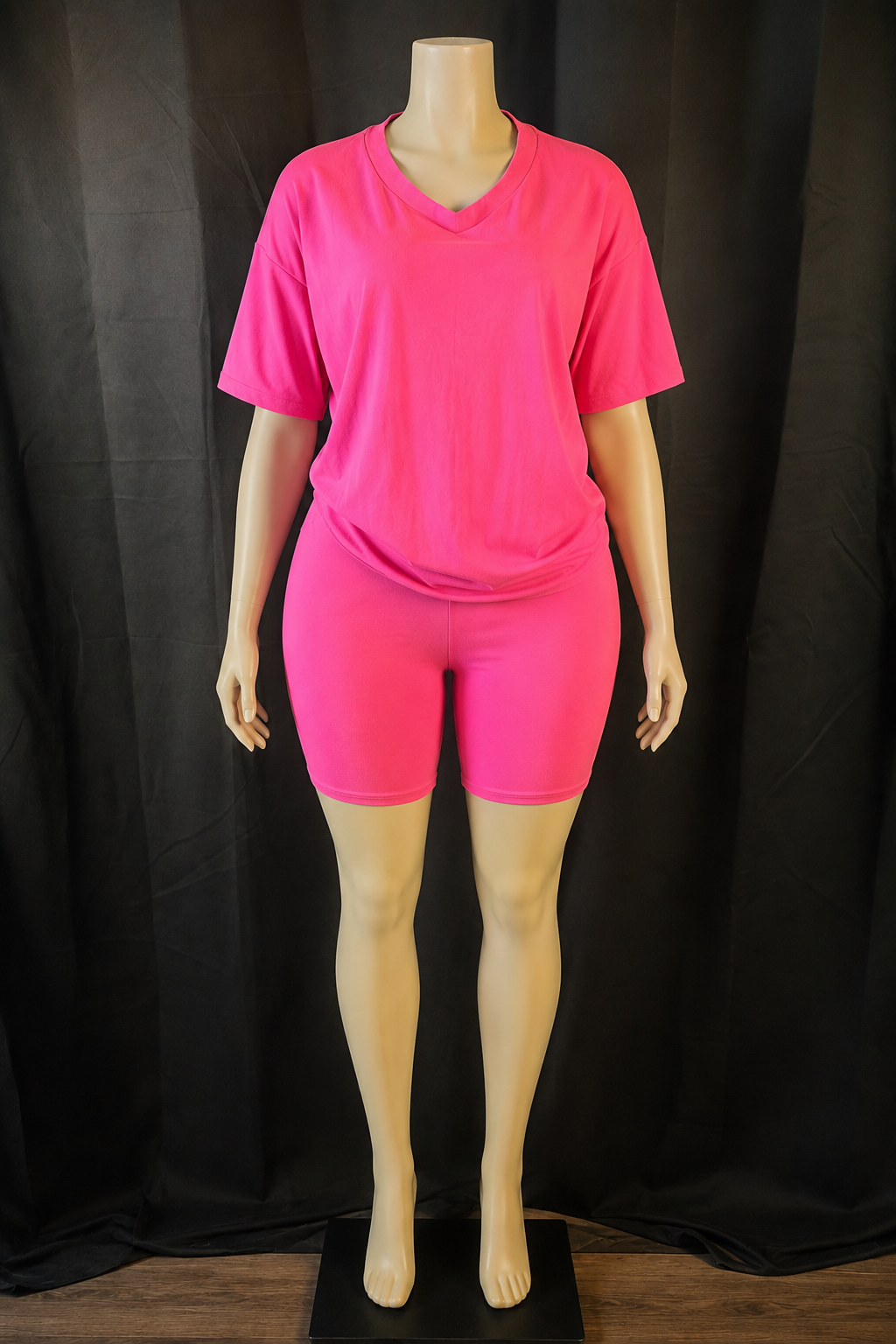 A mannequin wearing a bright neon pink short-sleeve V-neck t-shirt and matching fitted biker shorts, photographed against a black draped backdrop.