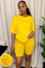 A woman modeling a bright yellow two-piece lounge set consisting of a short-sleeve top and matching fitted biker shorts, standing in front of a white curtain backdrop with a plant and a handbag visible.