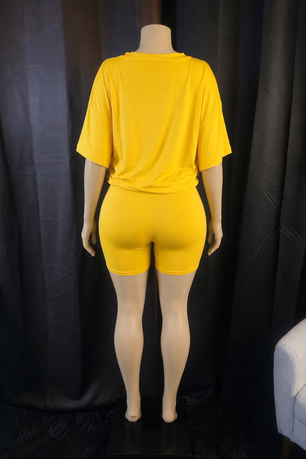 A bright yellow two-piece matching set consisting of a short-sleeve t-shirt and matching fitted shorts, displayed on a mannequin from the back against a black curtain backdrop.