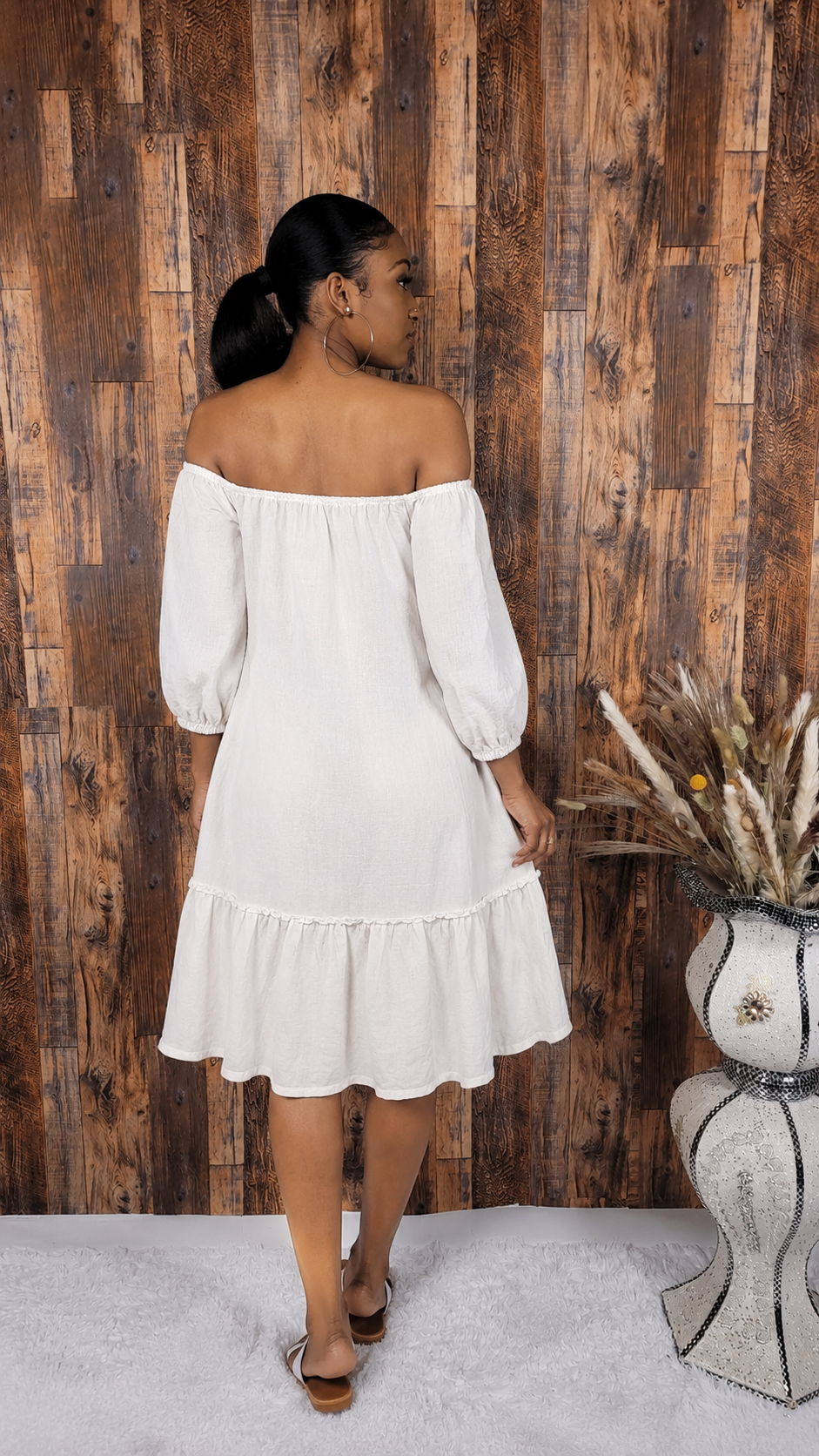 Woman in a white off-shoulder dress standing against a wooden wall with a decorative vase.