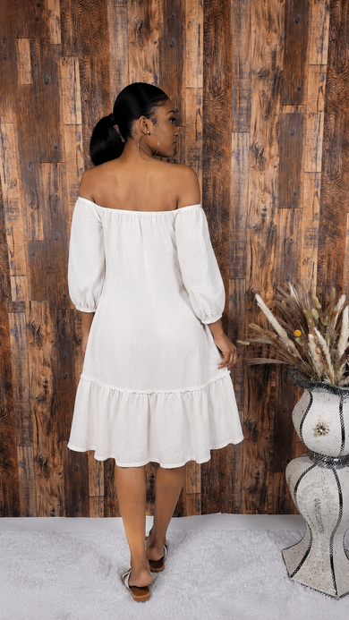 Woman in a white off-shoulder dress standing against a wooden wall with a decorative vase.